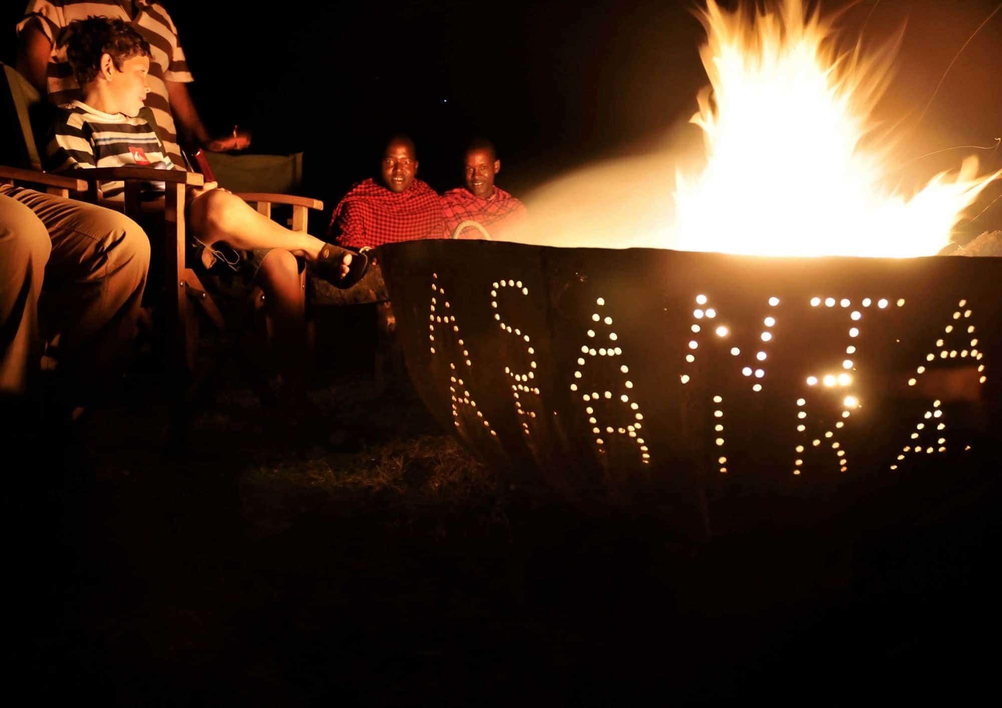 Guests and Maasai staff gathered around a glowing fire pit at night at a Serengeti safari camp, Tanzania