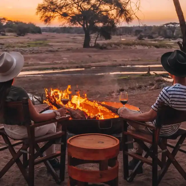 Couple with wine by a campfire at sunset overlooking a river valley on safari, Tanzania