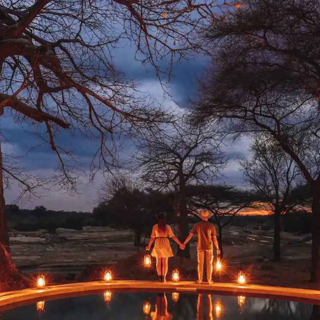 Couple holding hands by a lantern-lit plunge pool at dusk overlooking a river valley, Tanzania