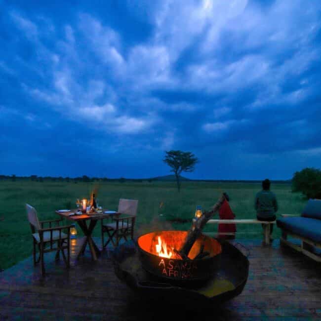 Fireside dining deck at dusk overlooking the Serengeti plains with a lone acacia, Tanzania