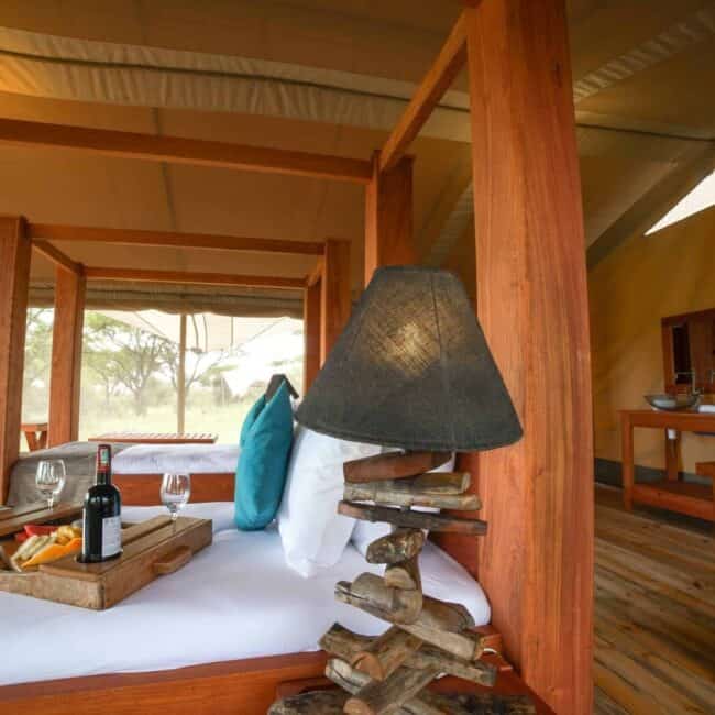 Luxury tent bed with wine tray and bush views through open mesh walls, Serengeti, Tanzania