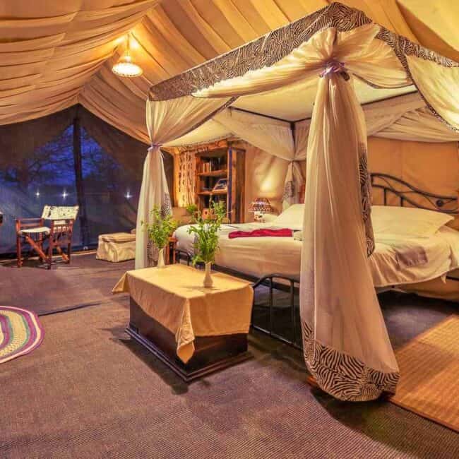 Canopied four-poster bed draped in mosquito netting inside a warmly lit luxury safari tent, Tanzania